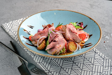 Warm beef salad featuring tender medium-rare slices, crispy potatoes, mixed lettuce, mustard seeds and balsamic reduction on rustic blue plate. Sophisticated gourmet main course