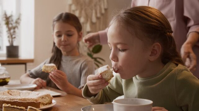 Chest up shot of little Caucasian girl enjoying delicious cake while sitting at table with little sister and drinking tea