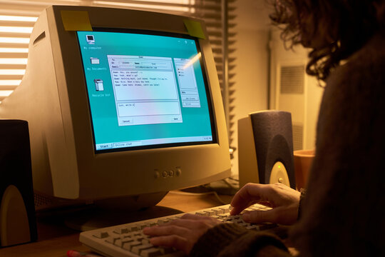 Caucasian young adult woman typing on vintage computer with CRT monitor, hands on keyboard, desktop speakers visible, chatting online in home office environment with window blinds in background