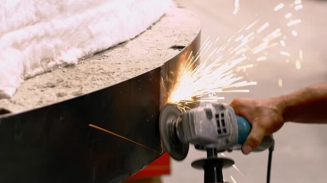 Close-up of worker using angle grinder on curved metal oven side producing bright sparks.