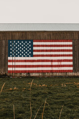 Large American flag painted on the side of a wooden barn. Grass field in the foreground under a grey sky. © Darien