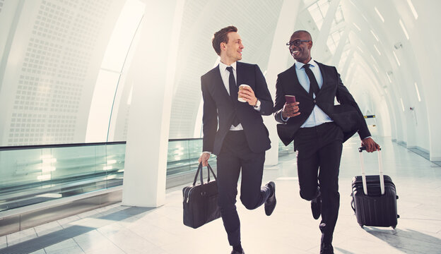 Smiling diverse busy businessmen running together through an airport for a flight. They are wearing suits and ties, with their luggage and passport in hand.