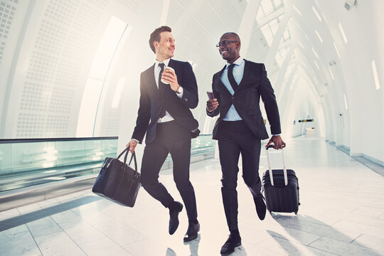 Two diverse smiling businessmen rushing through an airport for a flight. Their wearing suits and ties and have their luggage and passports in their hands.