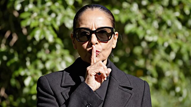 medium close up cinematic shot person facing camera performing silence gesture finger touching lips composed confident mood anonymous styling dark clothing sunglasses natural outdoor daylight green fo