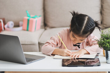 Fototapeta premium A cheerful Asian mother helps her young daughter do homework and color a book at home, while the father works on a laptop in the background. A cozy, bright, and modern family lifestyle.