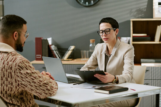 Middle aged Caucasian woman wearing glasses interviewing young adult man at desk, both sitting across from each other, woman holding digital tablet