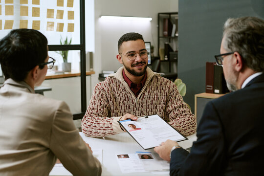 Young adult man sitting at desk interacting with two middle aged professionals during job interview, holding resume, making eye contact, engaging in conversation