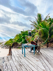 Fototapeta premium Relaxing day at a beachside table with palm trees and sea view. A person sits at a table on a beach deck among palm trees, looking towards the sea in the evening.