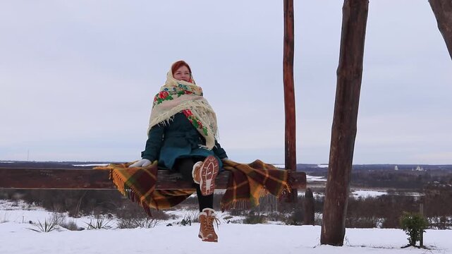 Beautiful girl swinging on a large swing with a view of a winter village and snowy fields