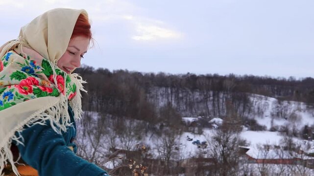 Beautiful girl with red hair in a traditional scarf blows snow off her hands Atmosphere of a happy winter day