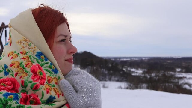 Close-up portrait of a young woman with red hair in a traditional headscarf looking at the winter landscape. Peace, cultural heritage, silence and inner harmony.