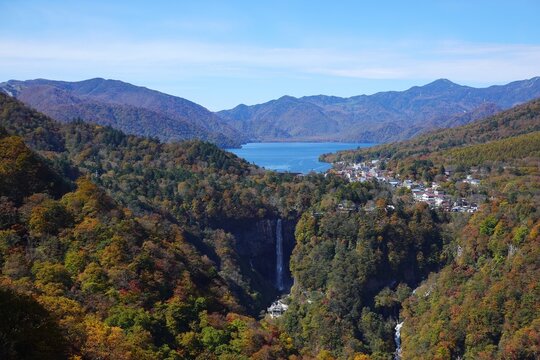 Panoramic aerial view of Kegon Falls and Lake Chuzenji with Chuzenji Onsen town during autumn peak in Nikko National Park, Tochigi, Japan. Vibrant red and orange foliage under clear blue sky.