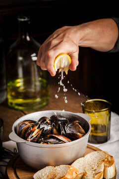 Hand squeezing fresh yellow lemon over silver metal pot filled with cooked black mussels seafood dish on wooden table with toasted bread slices and glass bottle of white wine in dark moody background.