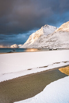 View of snow-covered beach meeting the turquoise sea beneath a moody sky, framed by majestic, snow-dusted mountains in the distance, Haukland beach Lofoten, Nordland, Norway.