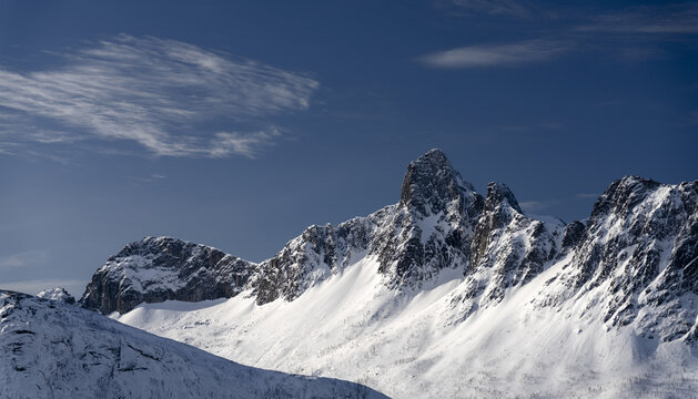 View of a stark, snow-laden mountain range under a crisp blue sky dotted with wispy clouds, creating a serene yet formidable winter landscape, Ornfjord Senja, Nordland, Norway.