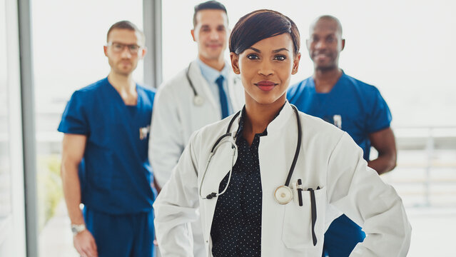 An empowered Black woman physician leads her diverse medical team. She makes direct eye contact with the camera while her colleagues work together in the background, symbolizing expertise and teamwork