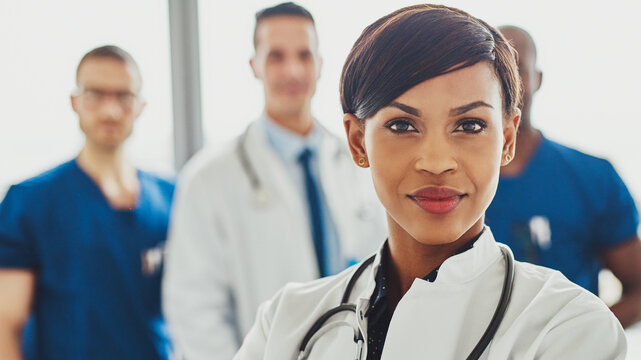 A black female doctor stands confidently in the foreground, looking directly into the lens. Behind her, a diverse medical team is visible, creating a sense of leadership and in a hospital setting.