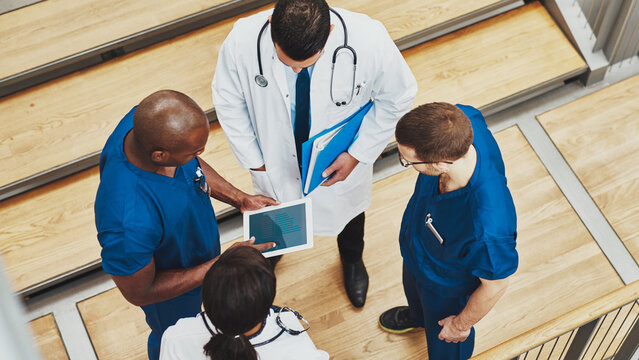 A top-down view of a diverse, multiracial team of four doctors gathered in a huddle. They are wearing blue surgical scrubs under white lab coats. Together, they are reviewing an iPad.
