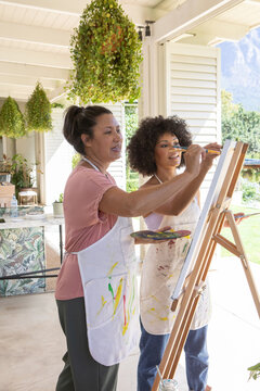 Asian instructor guiding her student painting on wooden easel at covered porch near plant
