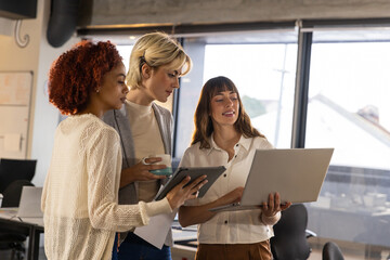 Diverse female team collaborating on laptop with tablet and mug in office by whiteboard