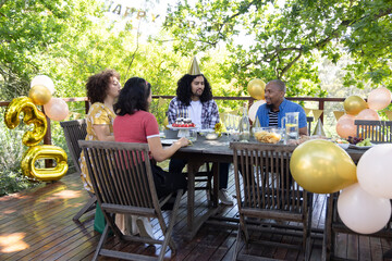 Friends celebrating birthday wearing party hats sitting around deck table with cake, balloons