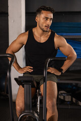 Obraz premium Muscular man pedaling stationary air bike in gym wearing black tank top green shorts and smartwatch
