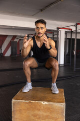Obraz premium Male in his 20s balancing in low squat on wooden plyometric box inside gym with smartwatch