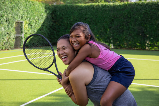 Asian mom carrying daughter piggyback on turf tennis court with boundary lines while holding racket