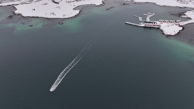 Aerial view of a boat cutting through the blue waters near the snow-covered Leknes harbor, creating a striking contrast, Leknes, Nordland, Norway.