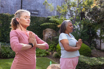 Fototapeta premium Diverse senior women friends meditating in garden wearing pink top and shirt with black watch