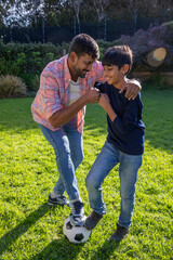 Asian father with son playing soccer and kicking ball on private backyard lawn under sunshine