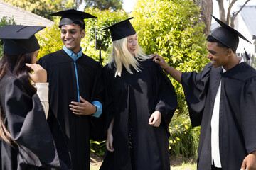 Diverse graduates standing on campus lawn by building wearing gowns, mortarboards, chatting