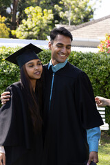 Indian friends standing in campus garden wearing graduation gowns, mortarboard near hedge © wavebreak3