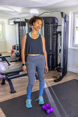 African American woman exercising with purple dumbbells on black mat in home gym with mirror wall