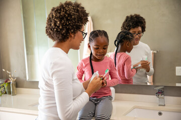 Diverse mother and daughter brushing teeth at home bathroom sink using toothbrushes