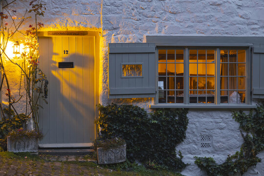 View of a quaint cottage facade glows with warm light spilling from the window and doorway onto the ivy-covered stone walls, Shaftesbury, United Kingdom.