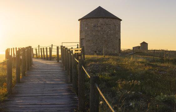 View of the old stone windmill stands majestically, bathed in the warm glow of the sunset, as a wooden walkway leads toward the horizon, Apulia, Braga, Portugal.