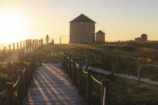 View of a wooden boardwalk leading toward ancient stone windmills silhouetted against a golden sunset sky in Apulia, Braga, Portugal.