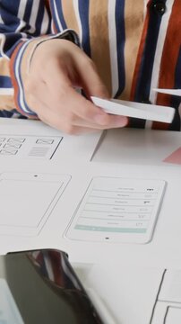 Vertical close-up of hands of unrecognizable female interface designer arranging low fidelity wireframes on desk, while working on user flow plan for mobile application