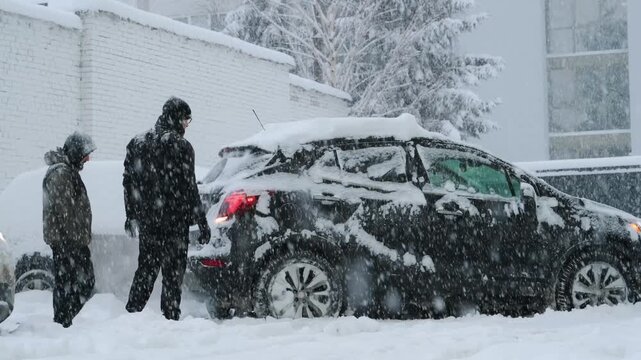 Man in a black winter coat is pushing a car stuck in deep snow during a snowfall with buildings and trees in the background creating a winter atmosphere
