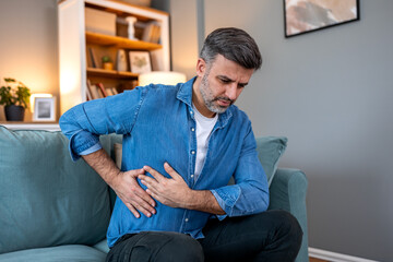 Side pain and kidney ache. Adult man sitting on sofa and holding painful side with hands. Healthcare and medicine concept. Handsome male suffering from liver, gallbladder or abdominal discomfort.