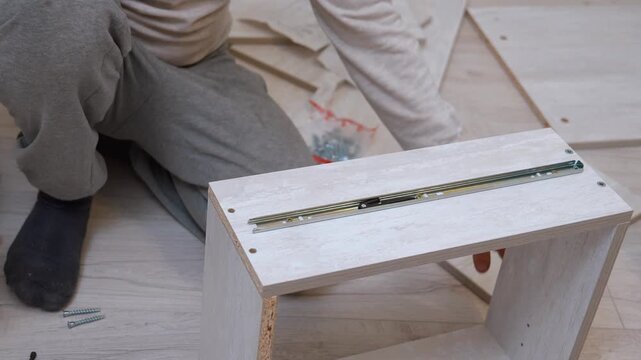 A young man assembles new wooden furniture on the floor using a cordless screwdriver or screwdriver, connects chipboard panels and installs drawer slides