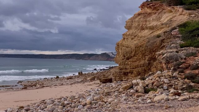 Atlantic waves roll onto a rugged pebble beach beneath sandstone cliffs at Zavial Beach, where clouds and raw textures shape a wild coastal atmosphere