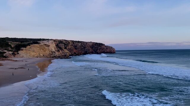 Coastal cliffs overlook ocean waves with surfers paddling at sunset Surfers paddle beneath dramatic cliffs at sunset near Zavial Beach, where Atlantic waves and fading light define the raw energy of t