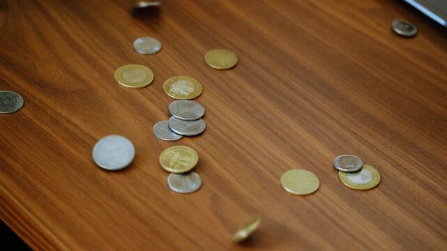 A close-up shot of a man&rsquo;s hand tossing small coins onto a wooden table surface. The coins scatter and slide naturally across the table. The action produces clean, crisp metallic coin sounds.