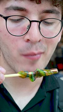 Izmir,Turkey,august 8, 2025.Young man with curly hair and glasses eating a colorful and sticky traditional ottoman candy, known as macun, on a wooden stick.He is savoring the sweet and sugary taste. 