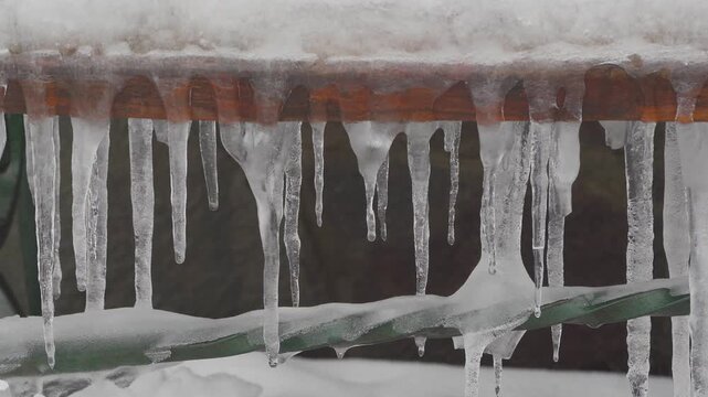 Melting icicles hanging from a  edge during spring thaw, A row of sharp ice formations hanging from a brown wooden beam covered with snow. High-quality stock footage of seasonal weather changes