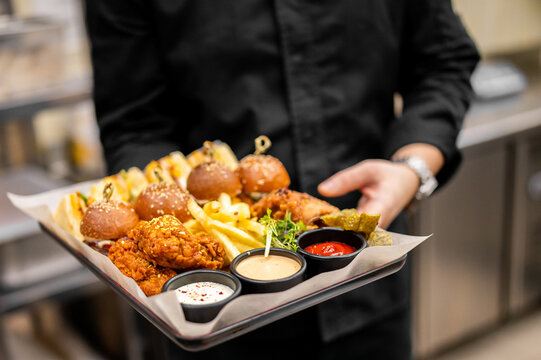 A close-up, horizontal, eye-level shot of a server holding a black tray laden with an appetizer platter of mini burgers, fried chicken, french fries, and dipping sauces.