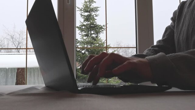 Man's hands working on a laptop at a table by the window on a snowy winter day. The scene captures the cozy focus and falling snow outside, 4K video