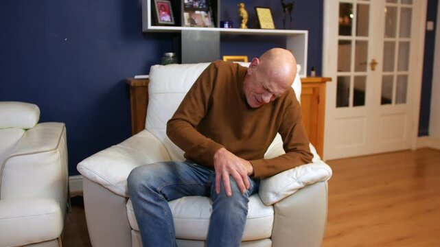 A man sitting on a white couch clutching his knee with a pained expression, suggesting physical discomfort. Living room setting with hardwood floors and visible home decor.
